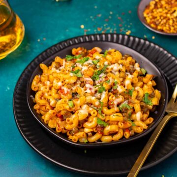 Masala macaroni topped with cheese, red pepper flakes and cilantro served in a black bowl