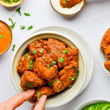 Butter chicken wings served in a white bowl along with extra sauce and condiments on the side and 2 hands grabbing a wing each.