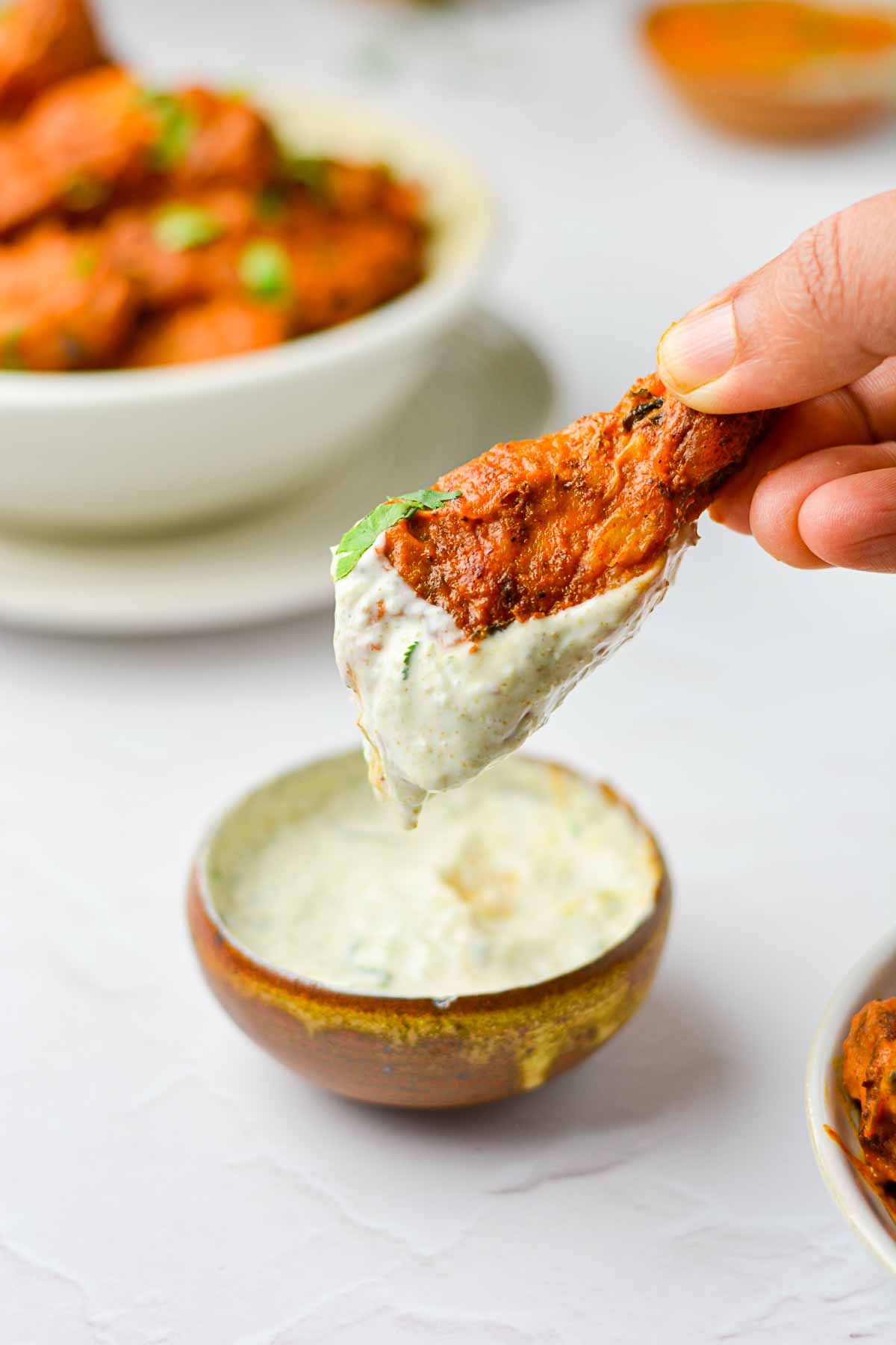 Butter chicken wing that was dipped into a creamy yogurt sauce being held up by a hand with a bowl of butter chicken wings in the background